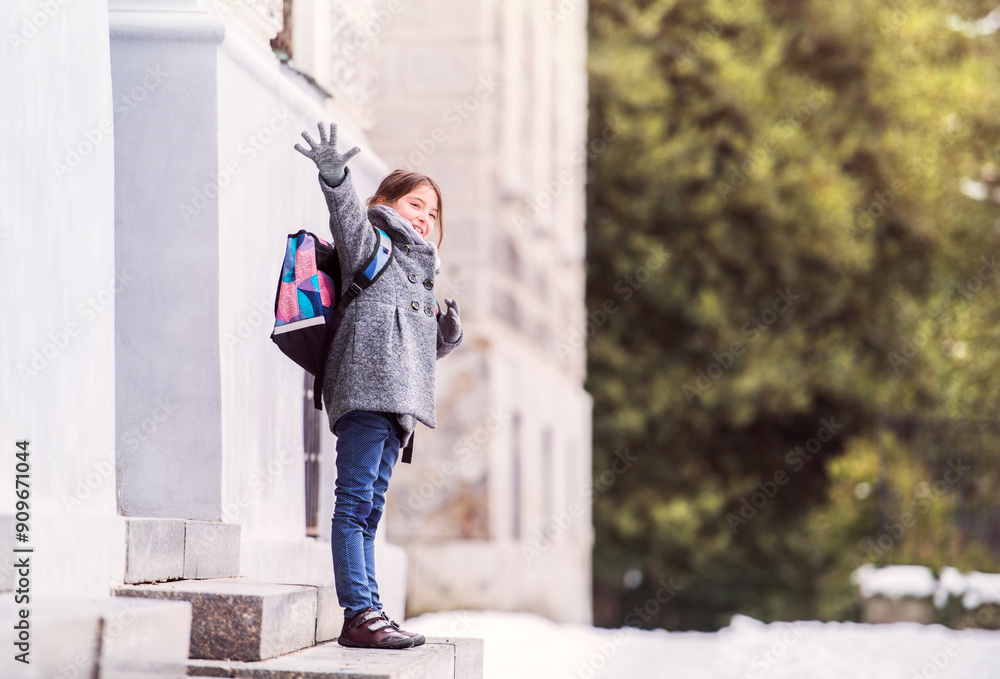 Portrait of schoolgirl in coat in front of school building, waiting for parents. Student girl with bag after day in elementary school. Portrait of schoolgirl in coat in front of school building, waiting for parents. Student girl with bag after day in elementary school.