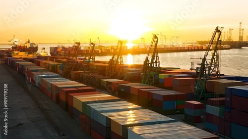 The sun rises over a bustling port with cargo ships alongside colorful containers. Silhouetted cranes stand tall against the glowing sky, emphasizing maritime trade.