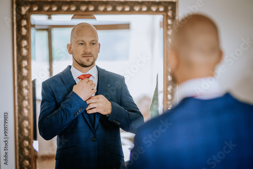 Cesis, Latvia - July 12, 2024 - A man in a blue suit adjusts his red tie while looking at himself in a mirror, preparing for a formal event. The room is softly lit. photo