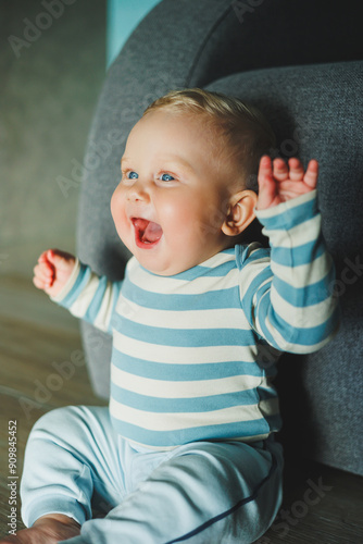 A blue-eyed baby with a smile on his face. A baby in a bodysuit and pants sits on the floor at home.