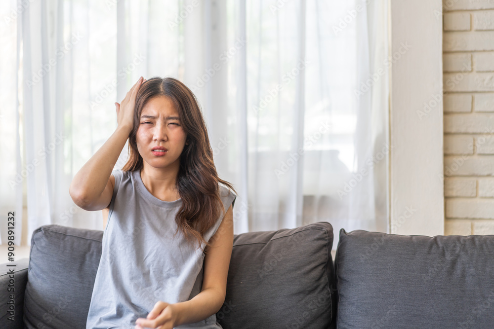 Young asian woman experiencing a headache sitting on a sofa with a pained expression, serious mood, health, wellness, stress, headaches and everyday struggles, pain and importance of self-care at home Young asian woman experiencing a headache sitting on a sofa with a pained expression, serious mood, health, wellness, stress, headaches and everyday struggles, pain and importance of self-care at home