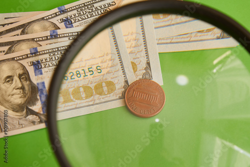 One cent coin on a background of hundred-dollar bills through a magnifying glass on a green background close-up, one cent on dollars, minimal profit, minimal wage, unprofitable bet. photo