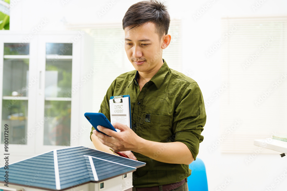 Asian businessman works alone at his desk, surrounded by a laptop, paperwork, tablet, and calculator, focusing on home and land mortgage agreements in a modern house model. Asian businessman works alone at his desk, surrounded by a laptop, paperwork, tablet, and calculator, focusing on home and land mortgage agreements in a modern house model.