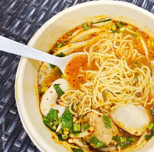 Close-up of a bowl of spicy noodle soup with fish balls, garnished with fresh green onions and herbs, served with a plastic spoon