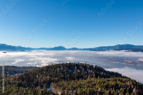 Panoramic view of snowcapped mountain peak Dobratsch in winter seen from tower Pyramidenkogel, Carinthia, Austria. Fog covered alpine valley surrounded by majestic Austrian Alps. Mystical atmosphere