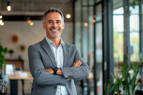 Smiling mature man in a grey suit standing with arms crossed in a modern office. Professional indoor portrait photography. Business and leadership concept