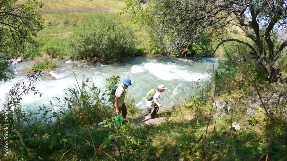 Two hikers are trekking through a gorge with a mountain river. A father and a boy are traveling in the summer in the mountains. Slow motion 2x Two hikers are trekking through a gorge with a mountain river. A father and a boy are traveling in the summer in the mountains. Slow motion 2x
