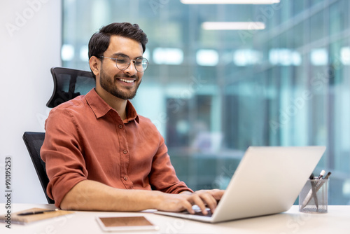 Fotografía Smiling man wearing glasses and an orange shirt working on laptop in modern office