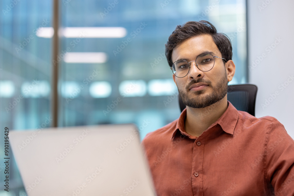 Serious businessman at modern office desk working on laptop. Young professional with glasses focused on completing project. Contemporary workspace with natural light and glass walls. Serious businessman at modern office desk working on laptop. Young professional with glasses focused on completing project. Contemporary workspace with natural light and glass walls.
