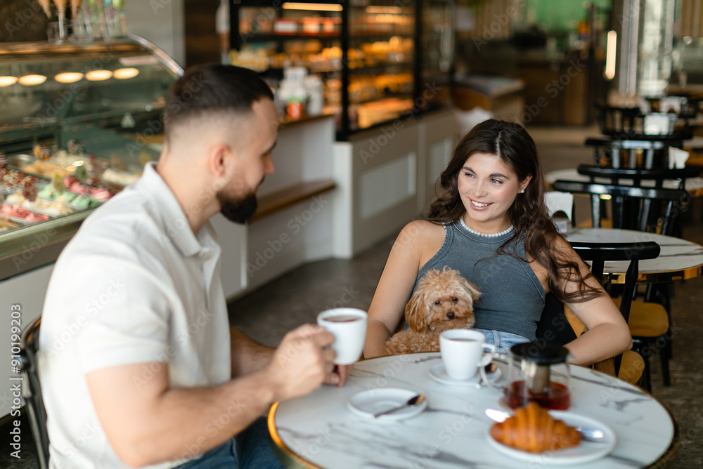 A woman with long hair with brown maltipoo and a bearded man in a cap are talking and eating croissants and drinking tea in cafe. Couple in love with little dog having breakfast in cafe. A woman with long hair with brown maltipoo and a bearded man in a cap are talking and eating croissants and drinking tea in cafe. Couple in love with little dog having breakfast in cafe.