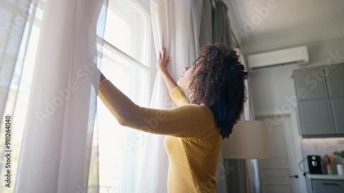 African American woman walking up to window and opening curtains in different sides. Curly female looking outdoor on street. Happily smiling while checking weather outside window. Peaceful day at home