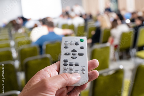 A hand is skillfully holding a remote control while in a conference setting with an engaged audience present