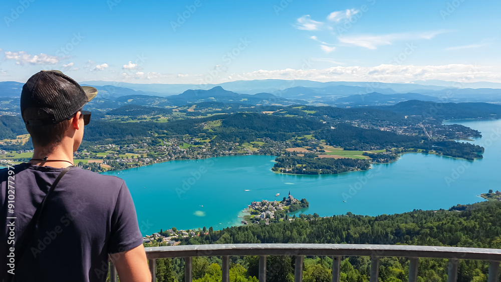 Tourist man with panoramic view of idyllic lake Woerthersee in summer seen from observation tower Pyramidenkogel, Carinthia, Austria. Lakeside villages surrounded by hills and mountains Austrian Alps Tourist man with panoramic view of idyllic lake Woerthersee in summer seen from observation tower Pyramidenkogel, Carinthia, Austria. Lakeside villages surrounded by hills and mountains Austrian Alps