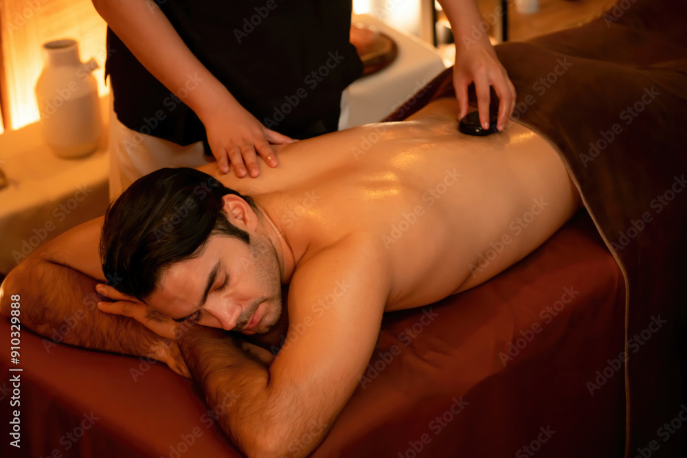 Obraz premium Hot stone massage at spa salon in luxury resort with warm candle light, blissful man customer enjoying spa basalt stone massage glide over body with soothing warmth. Quiescent