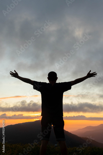 фотографія silhouette of young man standing on top of mountain peak with his hands raised above his head as symbol of worship and invocation of God's blessings with belief and  power of faith in God