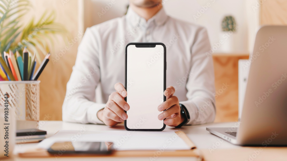 A business man holding a smartphone at a modern workspace filled with stationery and a laptop, perfect for tech and productivity themes. A business man holding a smartphone at a modern workspace filled with stationery and a laptop, perfect for tech and productivity themes.