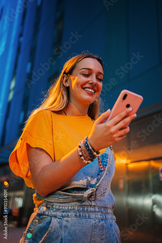 Vertical. one happy young girl using her smartphone to browse the internet while walking on the street at night. Female watching the social media with her cellphone. Girl typing on a mobile phone