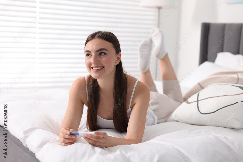 Happy woman holding pregnancy test on bed in room Happy woman holding pregnancy test on bed in room