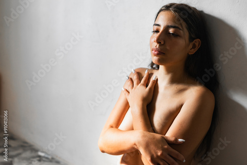 brunette latina woman covering her breasts with her hands folded while leaning against the wall with a sad look looking up.