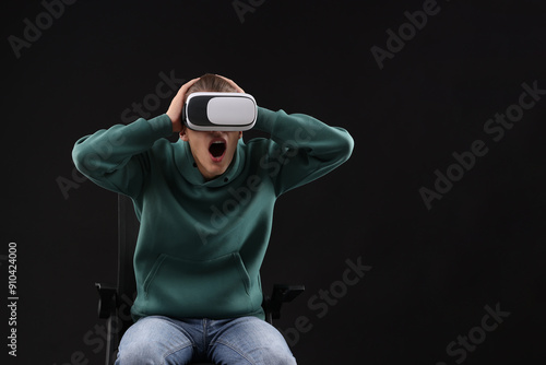 Emotional young man with virtual reality headset sitting on chair against black background, space for text