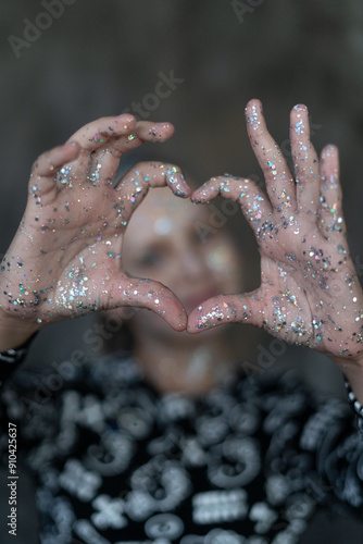 Selective focus on heart-shaped hands. Portrait of a girl with sequins on her face, smeared with silver sequins on her face, looking directly into the camera with a copy space