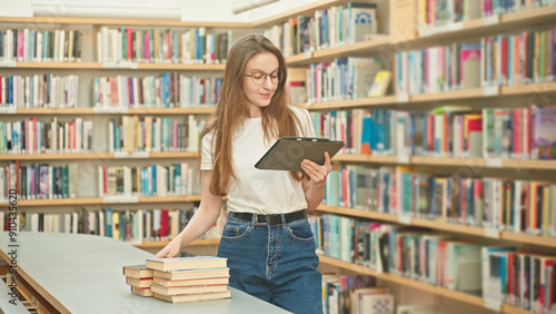 Caucasian woman wearing glasses with a tablet marking books in the library, working on a science project, doing homework for a tutor. Book selection, teacher prep, higher education, student e-learning