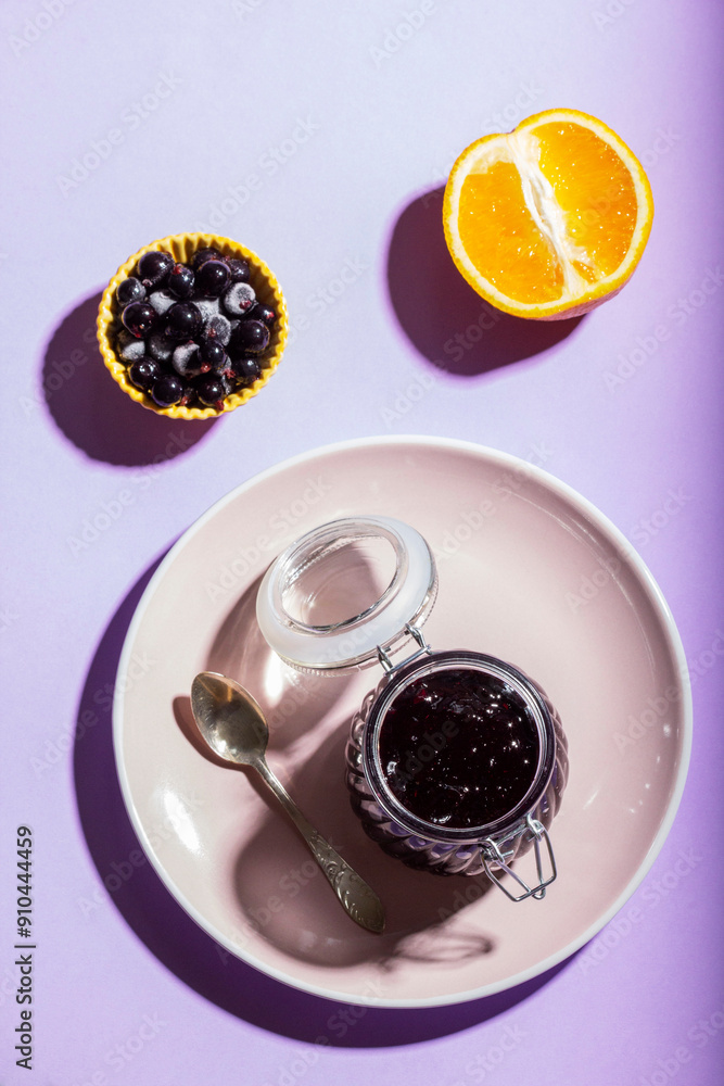 Blackcurrant jam with orange in a glass jar on a lilac background.