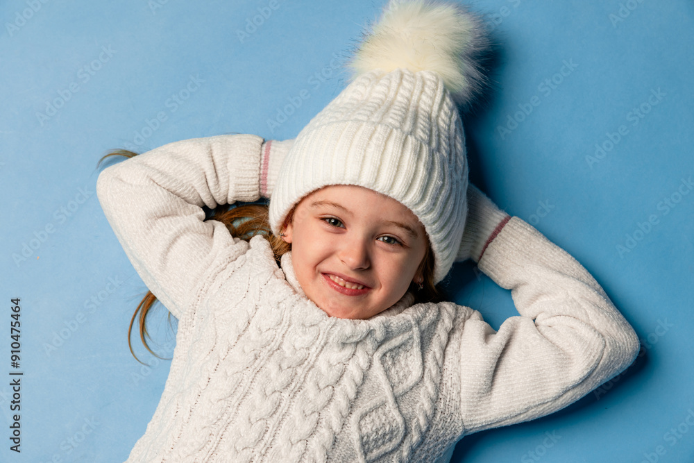 Top view child girl in a white sweatshirt and knitted hat with pompom on a blue background. children emotions, joy and delight. Advertising clothing and sales. Ski resort vacation, copy space Top view child girl in a white sweatshirt and knitted hat with pompom on a blue background. children emotions, joy and delight. Advertising clothing and sales. Ski resort vacation, copy space