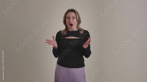 A young american or european excited and surprised blonde woman says wow, spreads hands on a beige studio background. An amazed and dumbfounded lady accepts unbelievable unexpected news, shows