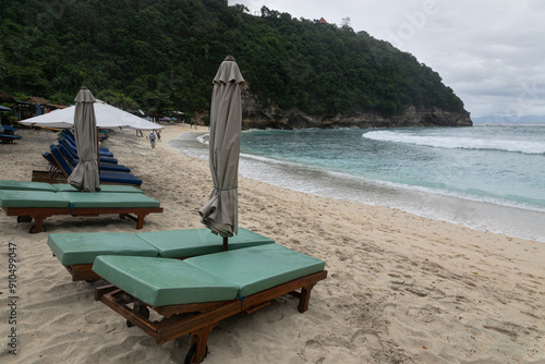 Row of empty deck chairs in cloudy weather on the Atuh beach on the island of Nusa Penida. Sandy beach, green islands,  sea surf and tropical nature around photo