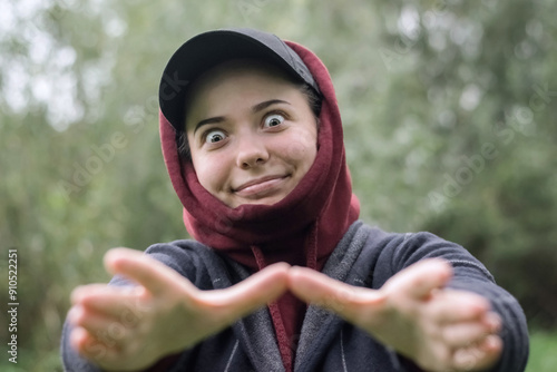 A funny girl in a cap and hood stretching out her arms with a strange smile and look in nature