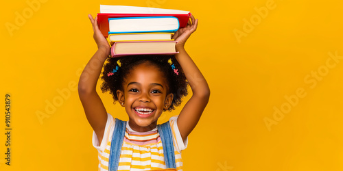 Cheerful young girl holding a pile of books over head head on yellow background. First day of school concept.