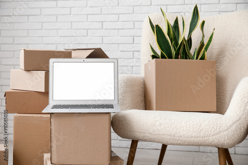 Blank laptop with moving boxes and armchair near white brick wall