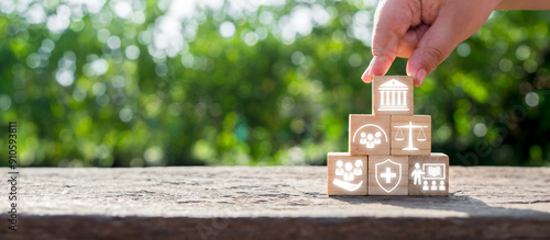 Obraz na płótnie Hand stacking wooden blocks with icons symbolizing social policy elements like healthcare, justice, and community support on a natural background