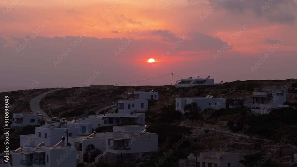 Drone rises above white homes on hillside of Donousa Greece as fire red ball of sun at sunset dips to hide below clouds on ocean