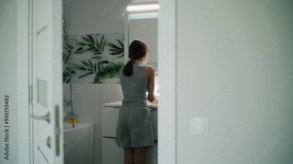 Young girl comes into bathroom, washes her face with cold water and looks at the mirror. Teenager stands in front of sink and uses electrical toothbrush to brush her teeth. Back view through door.