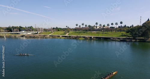 Drone flight crossing the Guadalquivir river (Seville) and where two canoes are training, crossing each other in the video shot. There is also a pedestrian bridge and a park with palm trees. photo