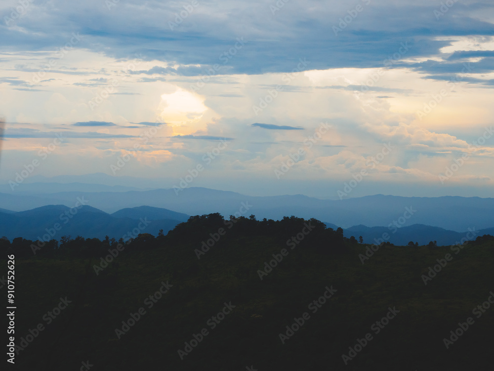Obraz premium Mountains under a cloudy sky at sunset. Lush greenery in the foreground contrasts beautifully with the misty distant mountains. Serene view of Phu Soi Dao National Park in Thailand.