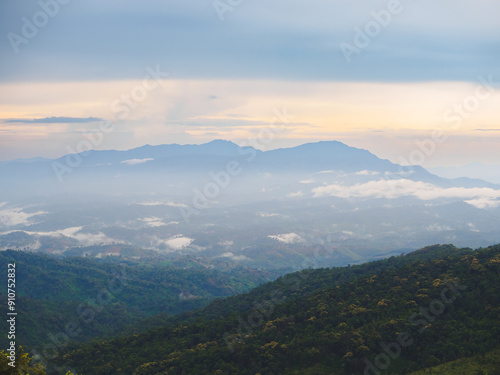 Mountains under a cloudy sky at sunset. Lush greenery in the foreground contrasts beautifully with the misty distant mountains. Serene view of Phu Soi Dao National Park in Thailand.