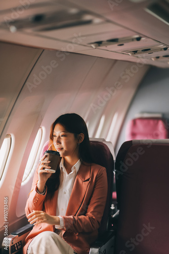 Portrait of a successful Asian businesswoman or entrepreneur in a formal suit on an airplane sitting in business class using a phone, computer laptop. Travel in style, work with grace.