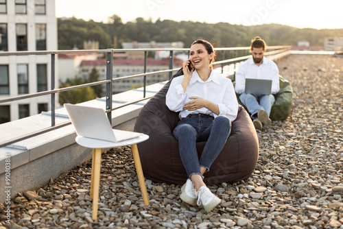 Colleagues working remotely on rooftop during sunny day. Smiling woman talking phone while sitting bean bag. Man focused on laptop in background. Modern open-air workspace.