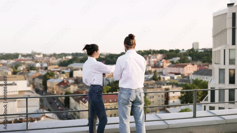 Business colleagues standing on rooftop, discussing project using technology devices. Urban cityscape view in background provides professional setting. Teamwork and collaboration concepts represented.