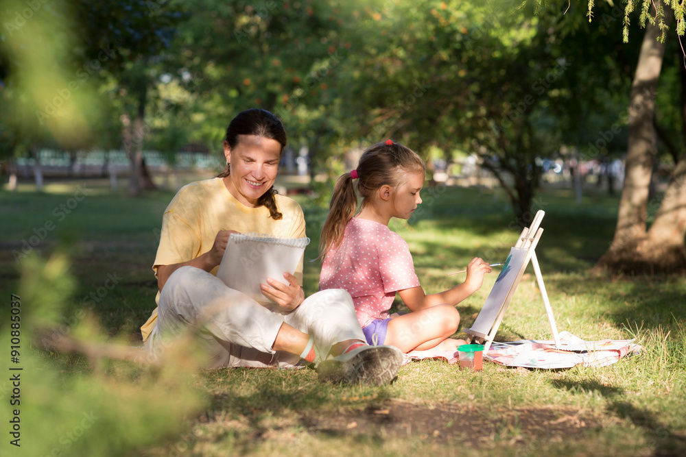 Fototapeta premium Happy family mom and child daughter having fun painting with wooden easel and doing picnic outdoor at city park on the green grass in summer day. KnowledgeForAll. Happy childhood.
