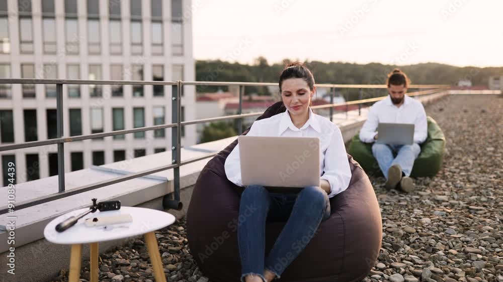 Two business people working outdoors on rooftop with laptops. Casual work environment with bean bag chairs and scenic city view. Focus on relaxation and productivity. Two business people working outdoors on rooftop with laptops. Casual work environment with bean bag chairs and scenic city view. Focus on relaxation and productivity.