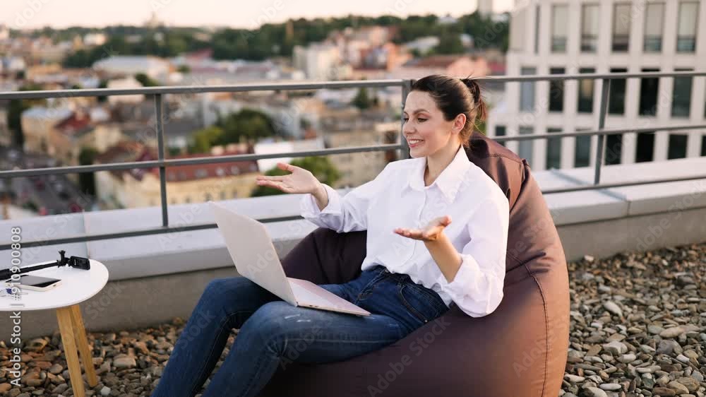 Smiling woman working on laptop outdoors on rooftop terrace, sitting on bean bag, enjoying fresh air, urban background, casual attire with jeans and white shirt, relaxed work environment. Smiling woman working on laptop outdoors on rooftop terrace, sitting on bean bag, enjoying fresh air, urban background, casual attire with jeans and white shirt, relaxed work environment.