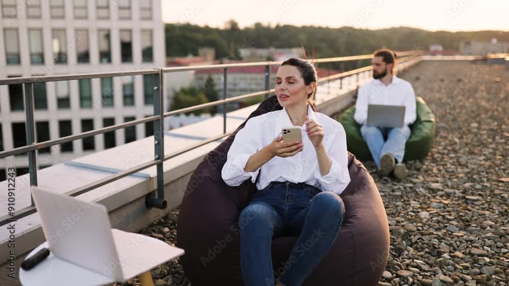 Casual work environment with employees using technology while sitting on bean bags outdoors on rooftop. Woman smiling with smartphone. Man focused on laptop. Modern office building in background. Casual work environment with employees using technology while sitting on bean bags outdoors on rooftop. Woman smiling with smartphone. Man focused on laptop. Modern office building in background.