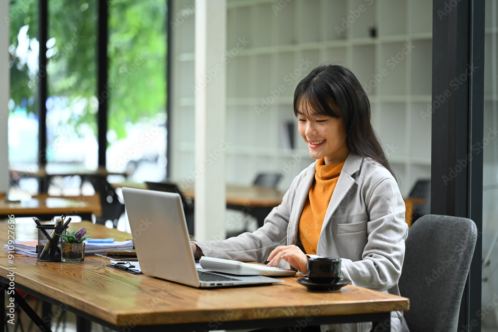 Asian businesswoman sitting at desk with laptop, using a calculator and analyzing financial documents Asian businesswoman sitting at desk with laptop, using a calculator and analyzing financial documents