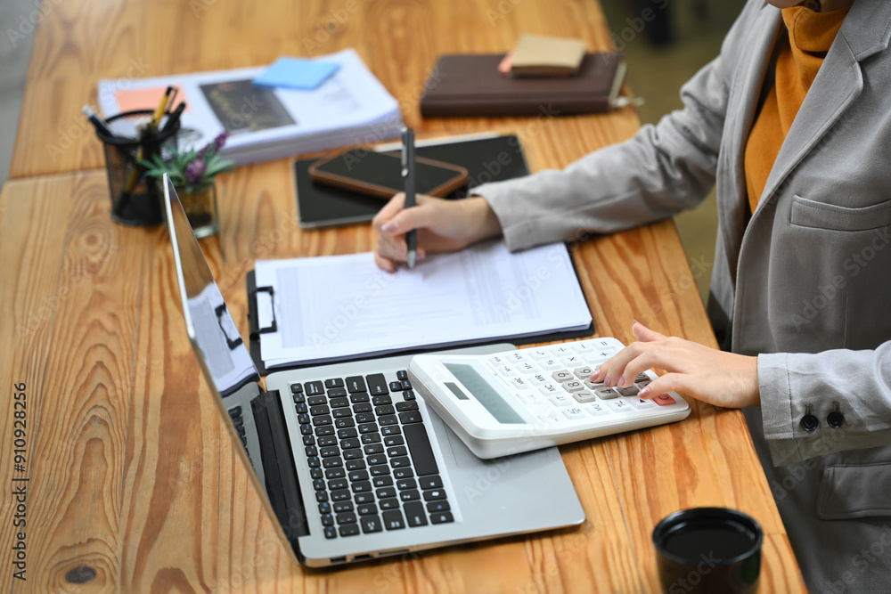Asian businesswoman sitting at desk with laptop, using a calculator and analyzing financial documents Asian businesswoman sitting at desk with laptop, using a calculator and analyzing financial documents
