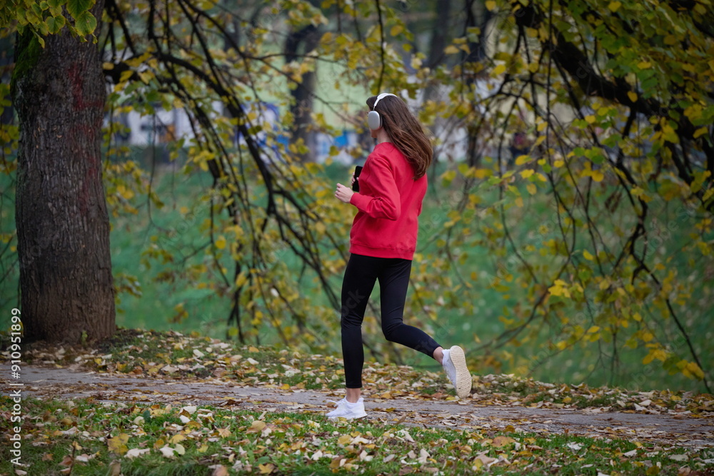 Fototapeta premium Young beautiful woman running in autumn park and listening to music with headphones on smartphone