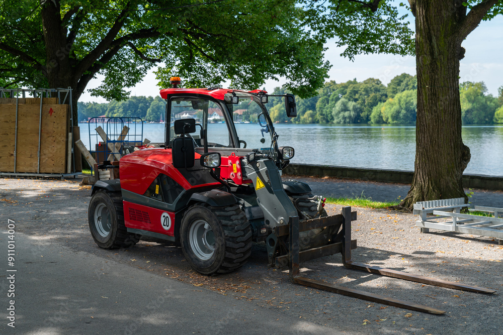 A red compact loader is parked by the riverbank, surrounded by green trees, enjoying a bright sunny day with clear skies. A red compact loader is parked by the riverbank, surrounded by green trees, enjoying a bright sunny day with clear skies.